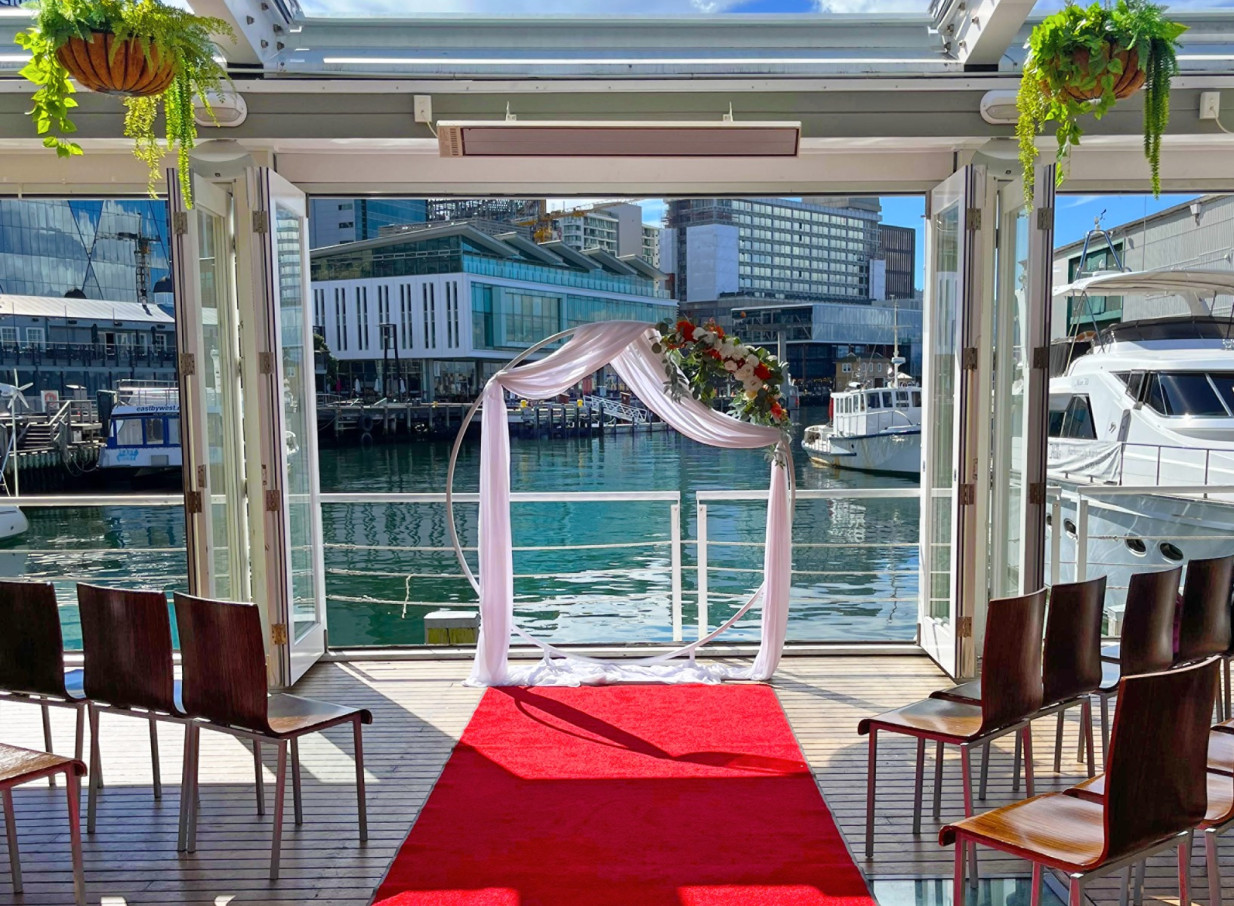 An elegant waterfront wedding ceremony with a floral arch and red carpet on Dockside, Wellington's historic open-air deck.