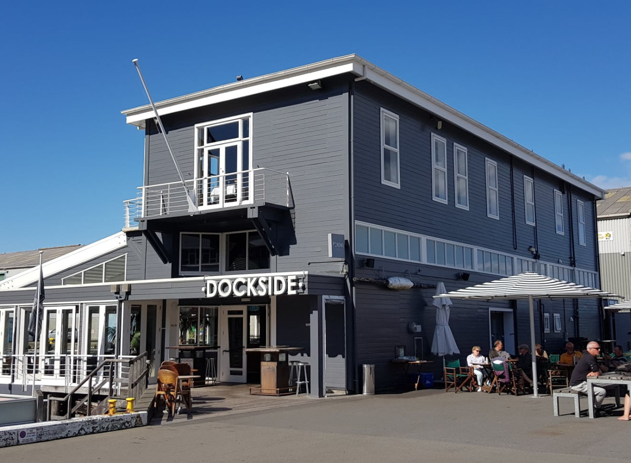 Dockside, a historic waterfront restaurant in Wellington, features a grey facade and a bustling outdoor dining area with striped umbrellas.