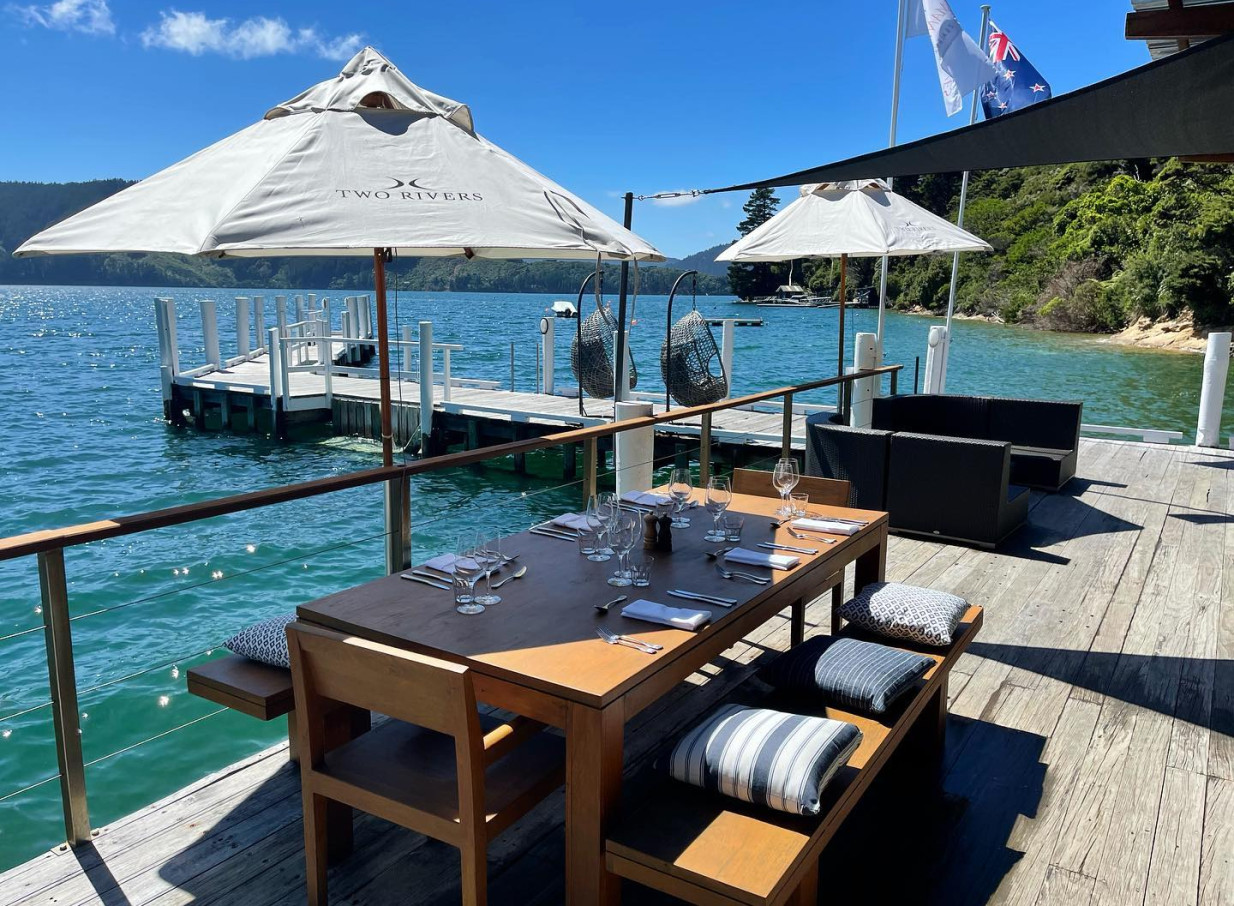 An outdoor waterside dining area on a wooden deck at Bay of Many Coves Resort, Marlborough, offering natural luxury with stunning bay views.