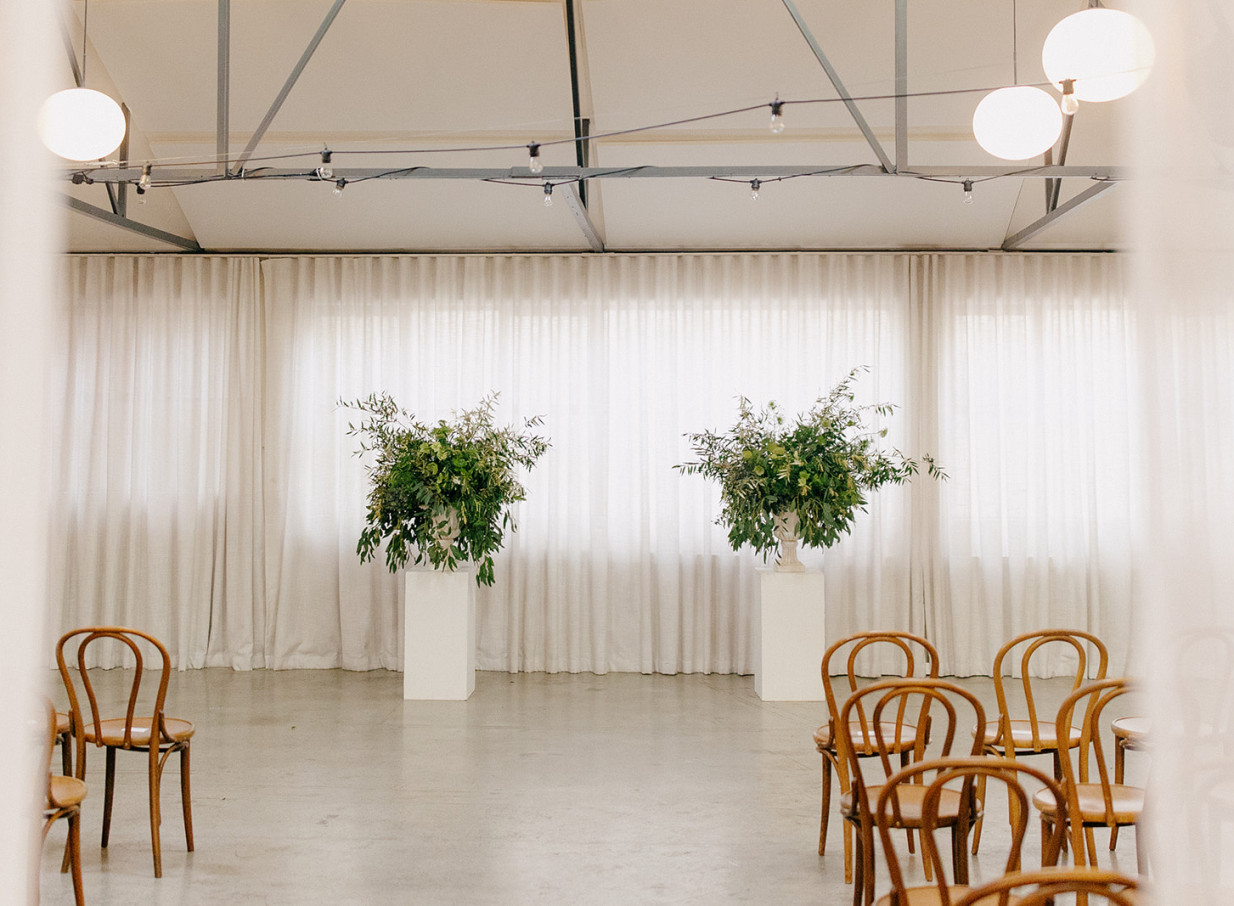 An industrial-chic ceremony at Whisky & Wood, Wellington, features sheer curtains, lush floral arrangements, and bentwood chairs.