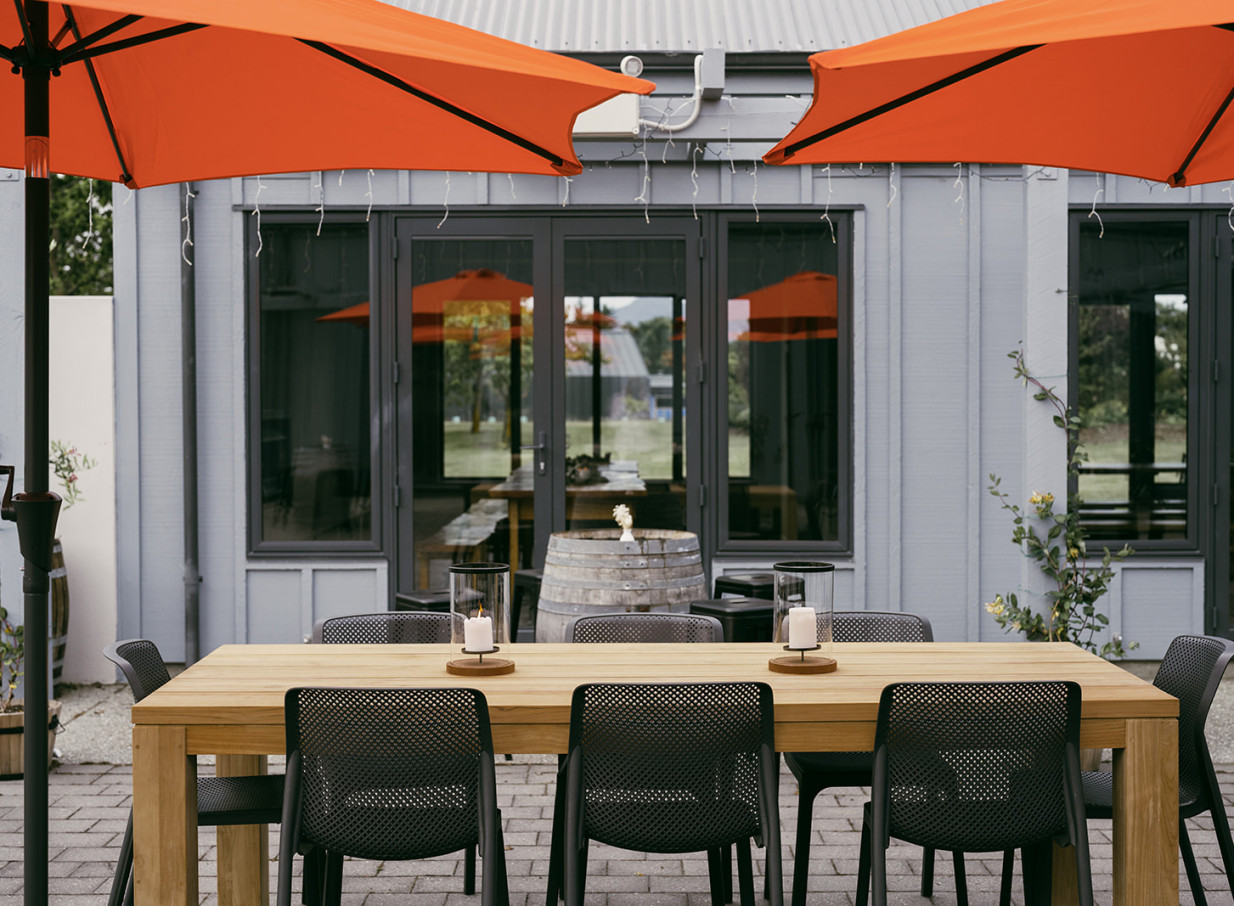 The Venue Wānaka's rustic-glam outdoor courtyard in Wanaka features a long wooden table, modern chairs, and bright orange umbrellas.