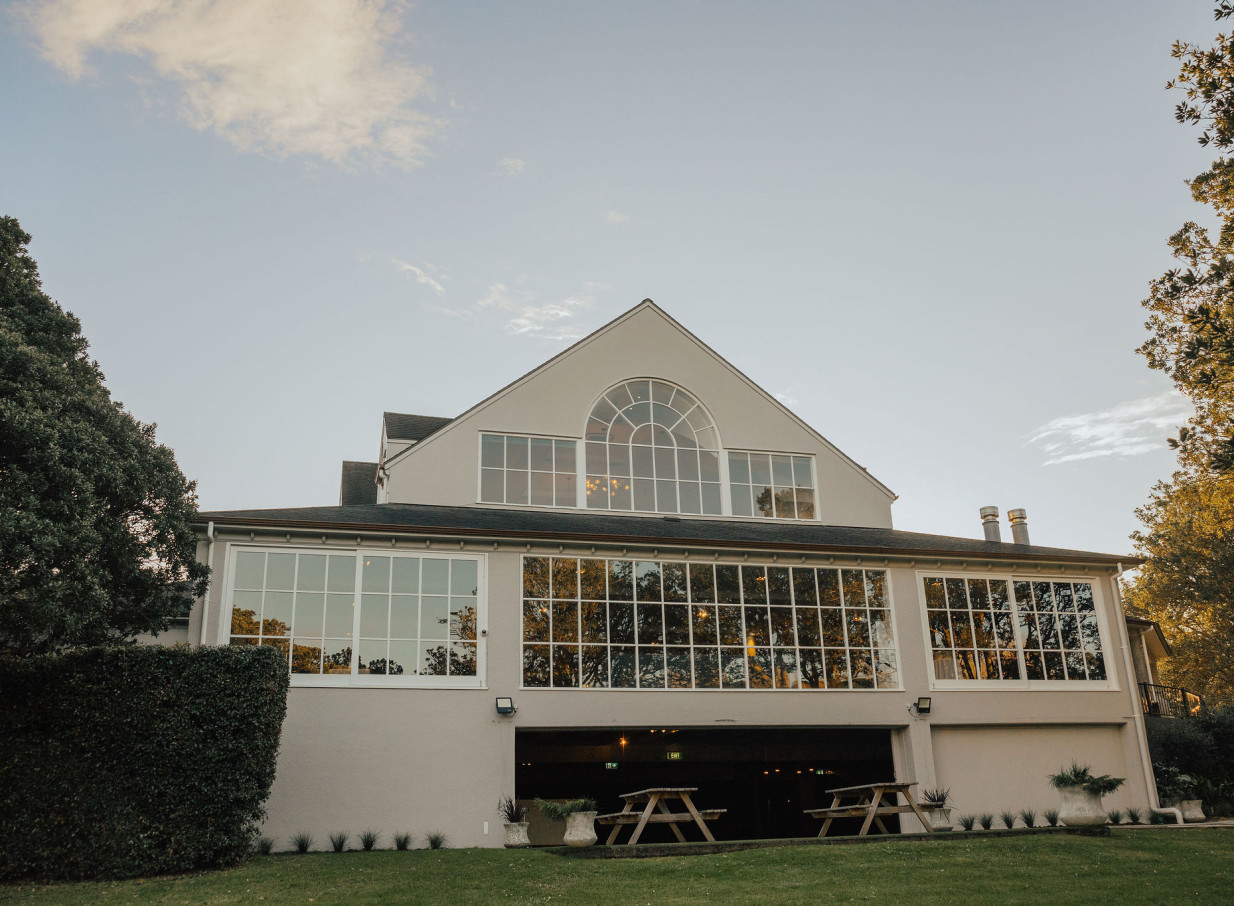 Orakei Bay, Auckland, presents its modern event space with a glass-fronted exterior, an arched window, and an inviting outdoor lawn.