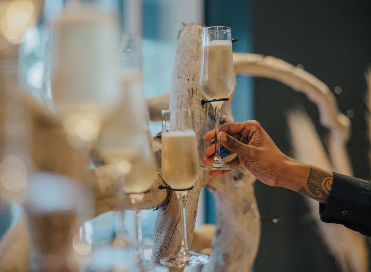 A hand holds a champagne flute amidst a natural wood display at the elegant waterside Orakei Bay venue in Auckland.