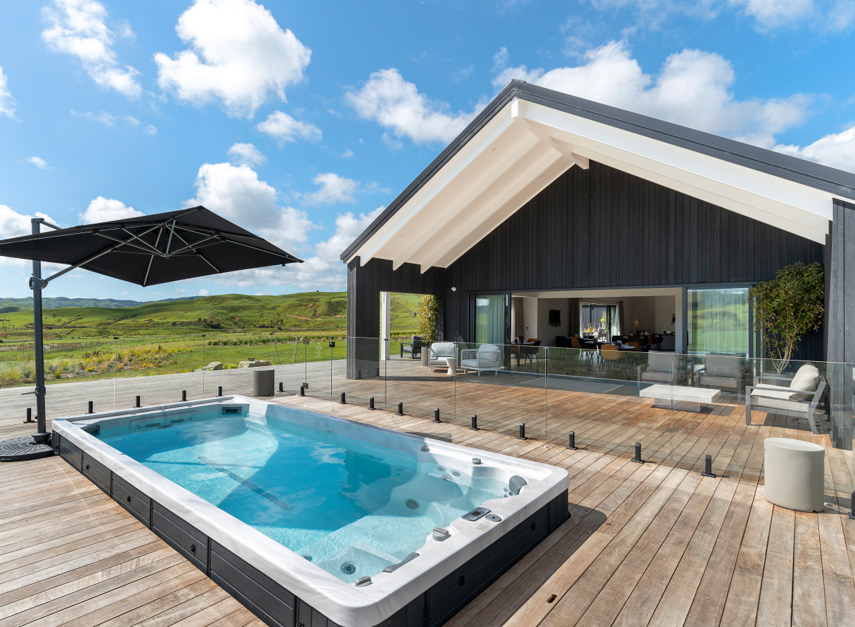 The modern Te Karaka Lodge in Waikato features an outdoor heated spa pool on a wooden deck, overlooking rolling hills.