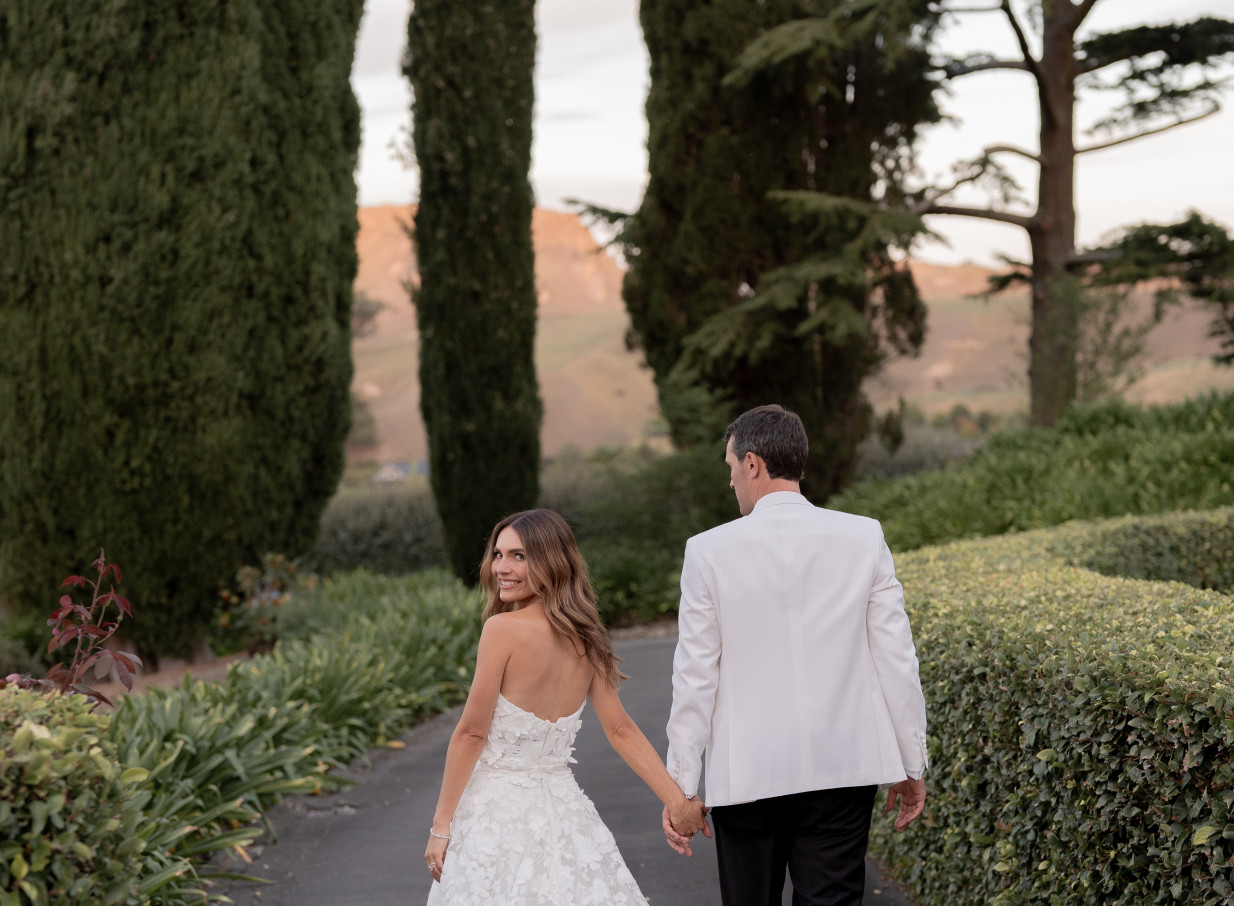An elegant wedding couple walks hand-in-hand along a tree-lined path at the 1920s Mediterranean Kōtare Estate in Hawke's Bay.