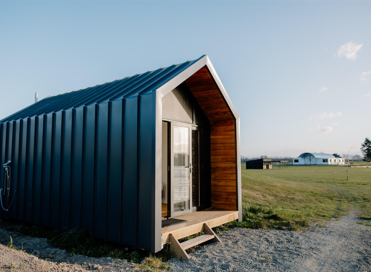 A contemporary lakeside cabin with dark cladding and a warm wooden entry deck at Backpaddock Lakes in Hawke's Bay.