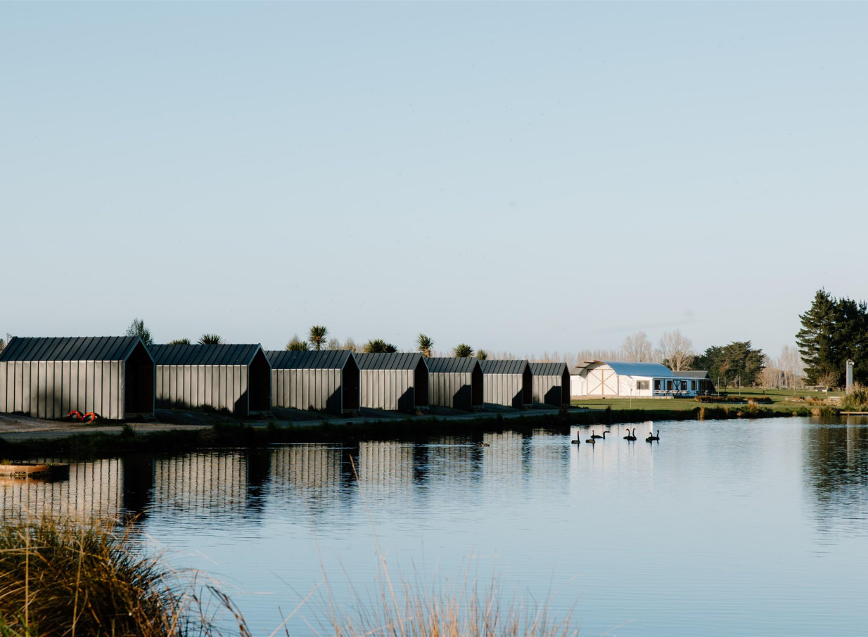 Seven contemporary-style cabins line the tranquil lake at Backpaddock Lakes, Hawke's Bay, with swans swimming nearby.