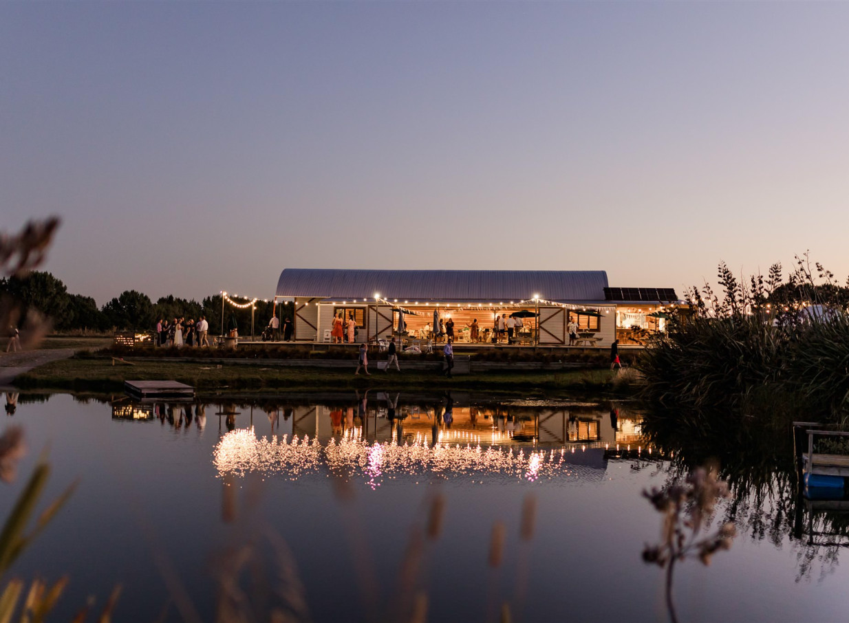 The rustic barn venue at Backpaddock Lakes, Hawke's Bay, glows during a festive evening reflected in the lake.