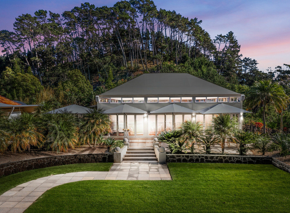 The elegant Spanish Mission-style homestead at Puketutu Island Estate in Auckland glows at dusk, surrounded by lush gardens.