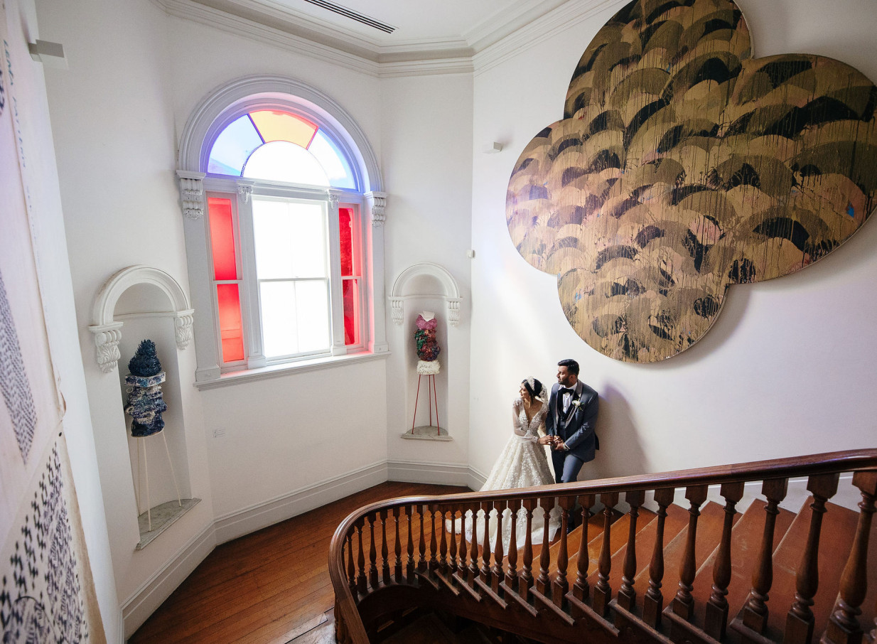 A couple on the grand staircase of Auckland's Italianate Pah Homestead, featuring an ornate window and contemporary artwork.
