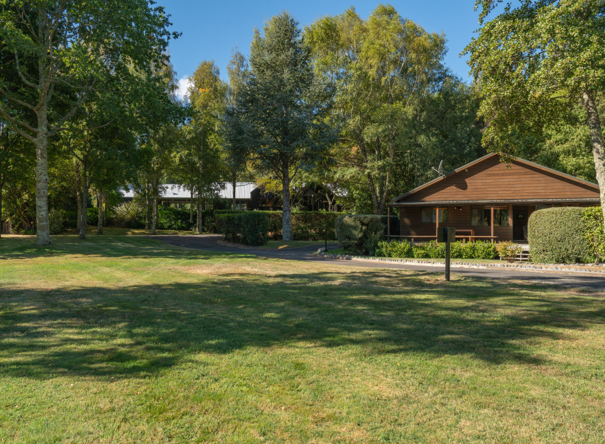 A rustic wooden chalet at Tongariro Lodge in Taupo is nestled amidst green trees and a sunlit lawn, reflecting its fishing lodge atmosphere.