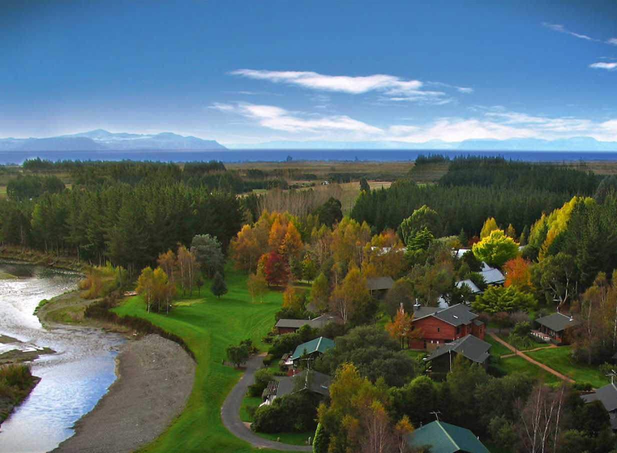 Aerial view of Tongariro Lodge's rustic chalets in Taupo, an iconic fishing lodge, surrounded by autumn trees, river, and Lake Taupo.