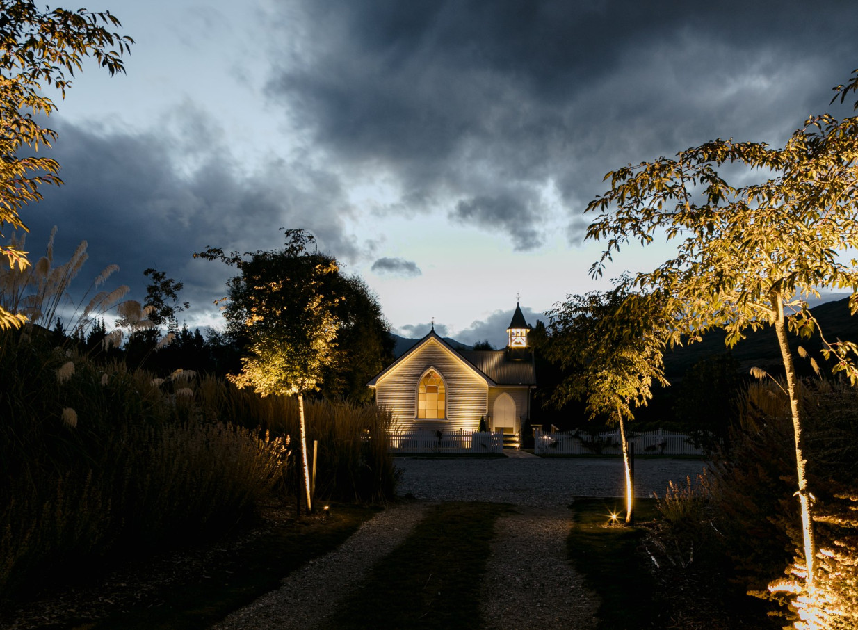 An illuminated, century-old heritage chapel, Waikākā Church in Queenstown, is framed by trees at dusk.
