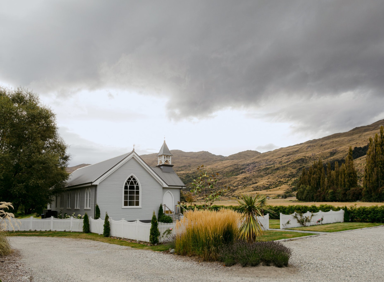 The historic Waikākā Church Queenstown, a restored chapel with a steeple, is set against mountains.