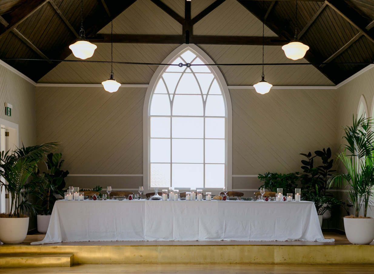 A dining setup within the historic Waikākā Church in Queenstown, featuring a high vaulted ceiling and a prominent Gothic arched window.