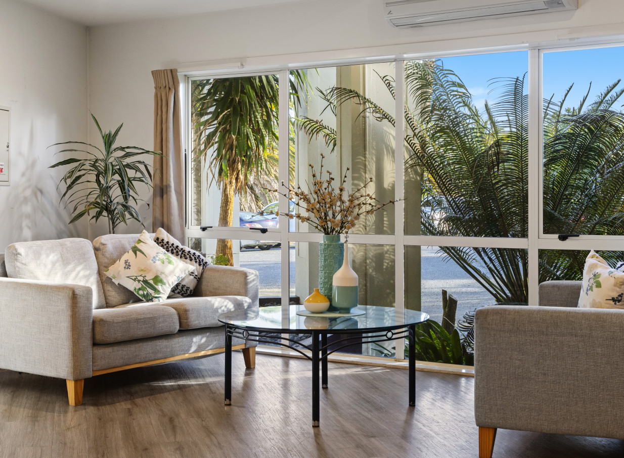 A modern living room at Taipa Beach Resort, Northland, features light grey sofas and tropical palm tree views.