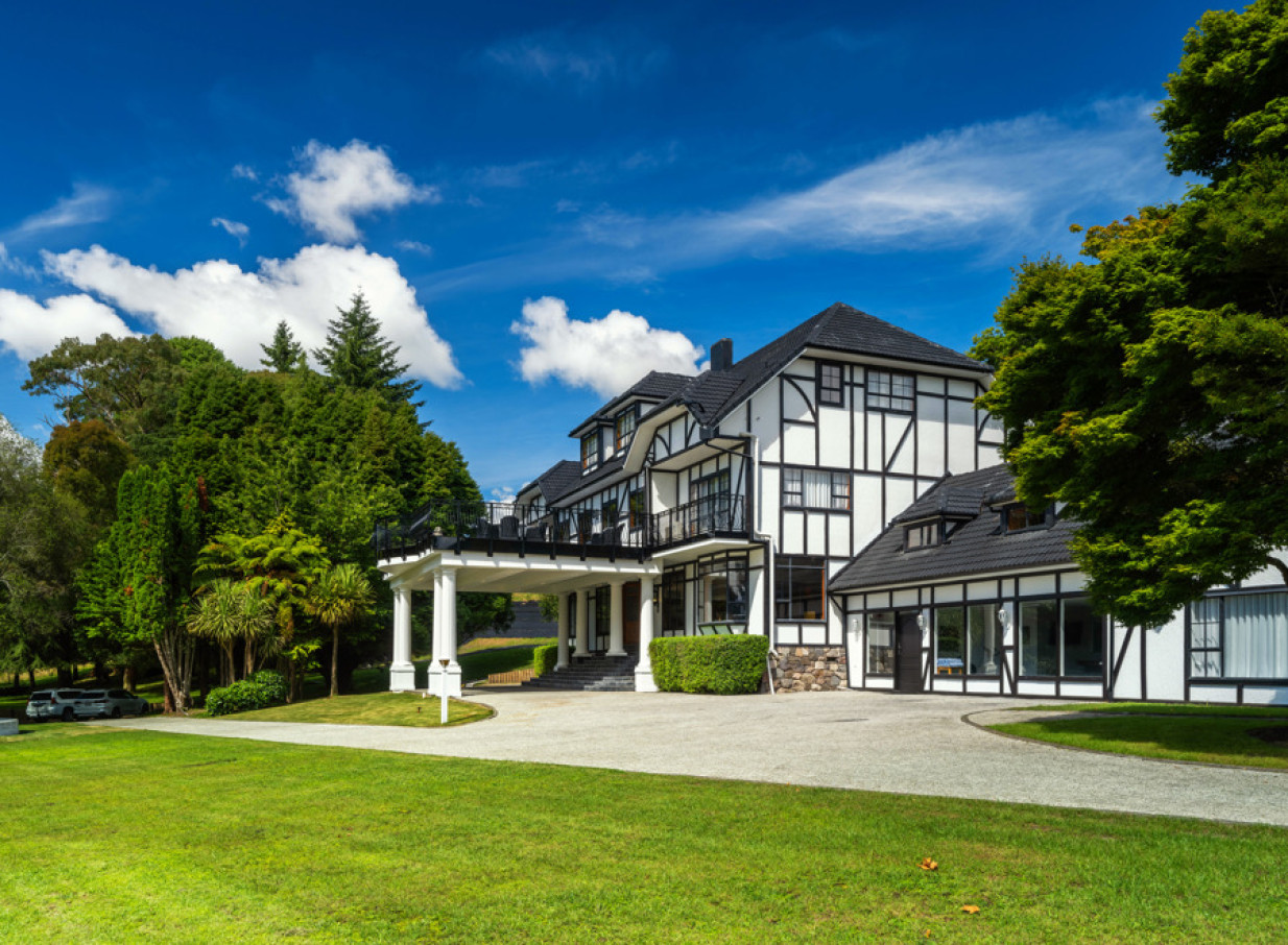 The elegant, Tudor-style Hana Lodge in Rotorua presents its grand entrance amidst a vibrant green landscape.