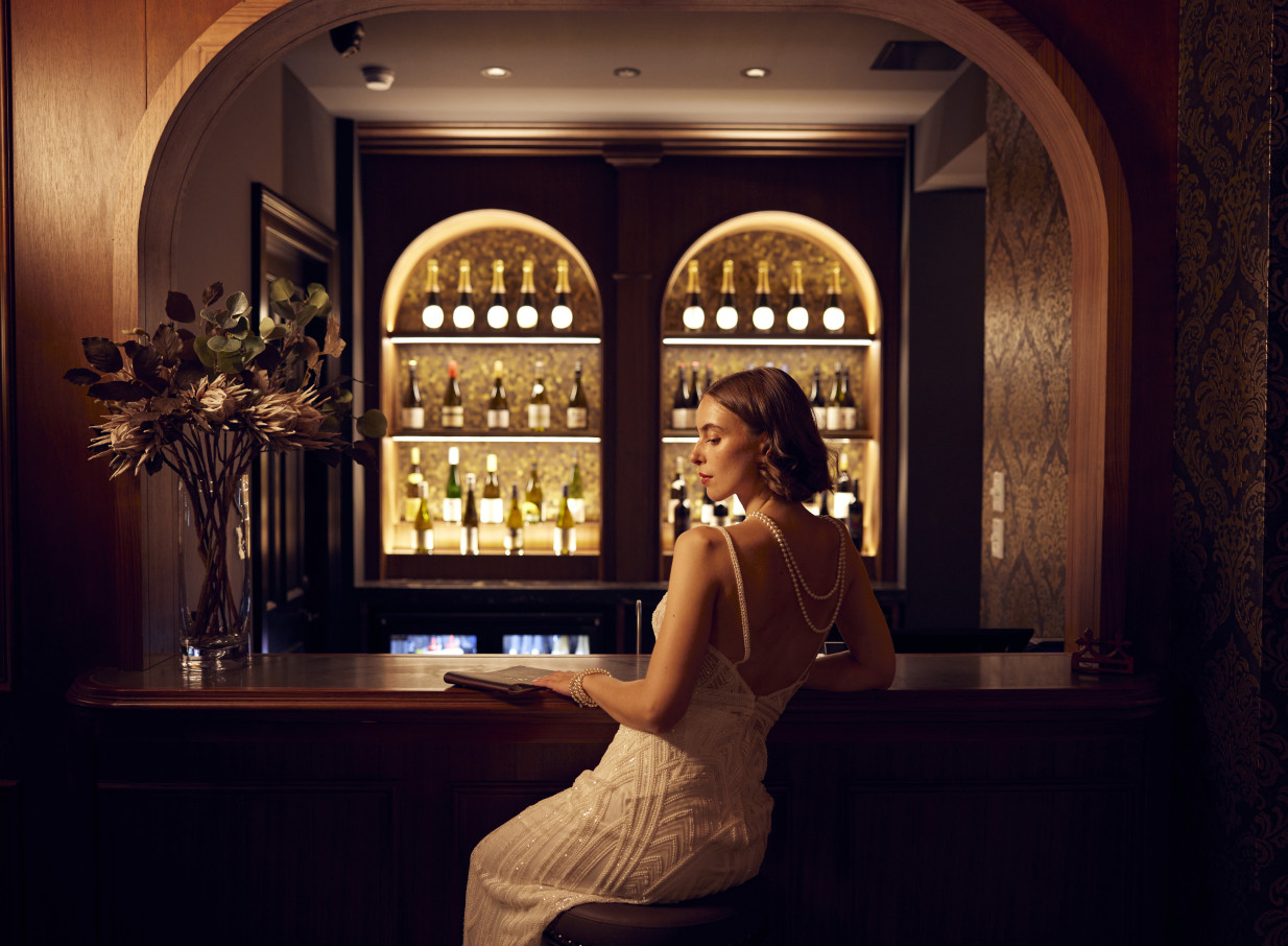An elegant woman enjoys the sophisticated speakeasy-inspired bar at The Clements Hotel, Waikato, reflecting its Beaux-Arts grandeur.