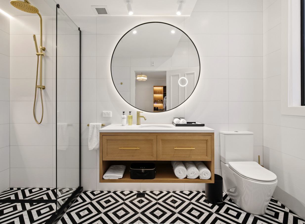 A contemporary bathroom at The Clements Hotel, Waikato, features geometric floor tiles, a wooden vanity, and a backlit circular mirror.