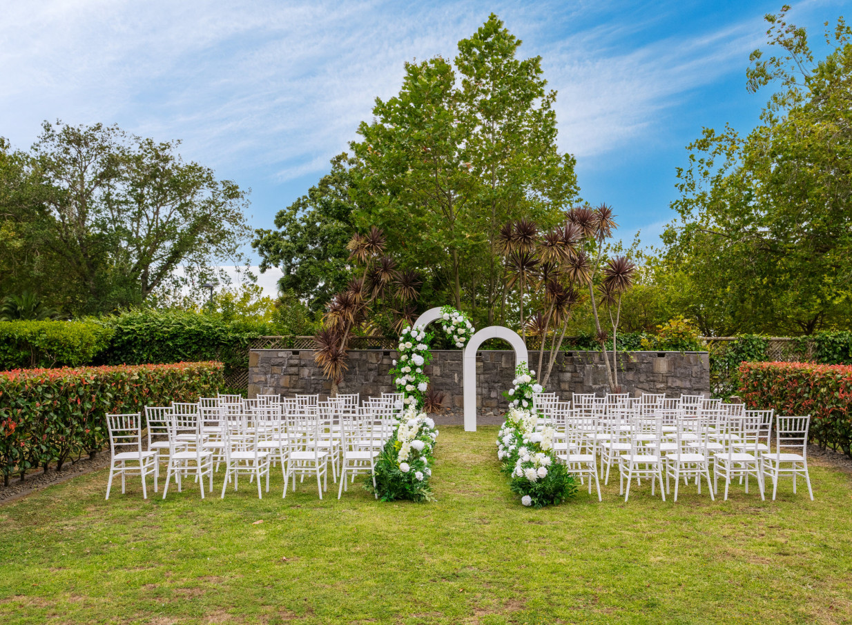 An outdoor wedding ceremony setup with white chairs and floral arches in the scenic gardens of Novotel Auckland Ellerslie.