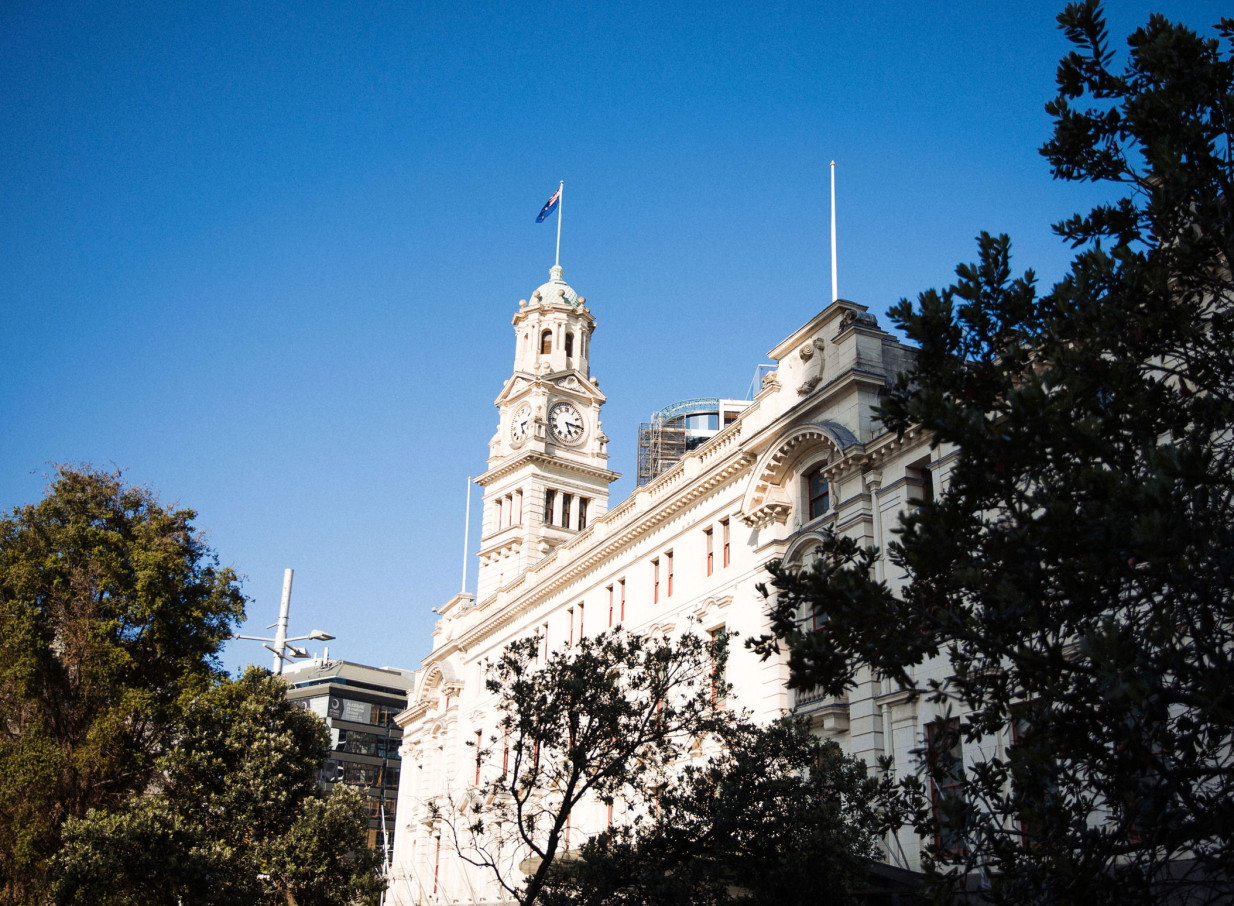 The grand, neo-Baroque Auckland Town Hall, with its iconic clock tower, is framed by trees against a clear blue sky.