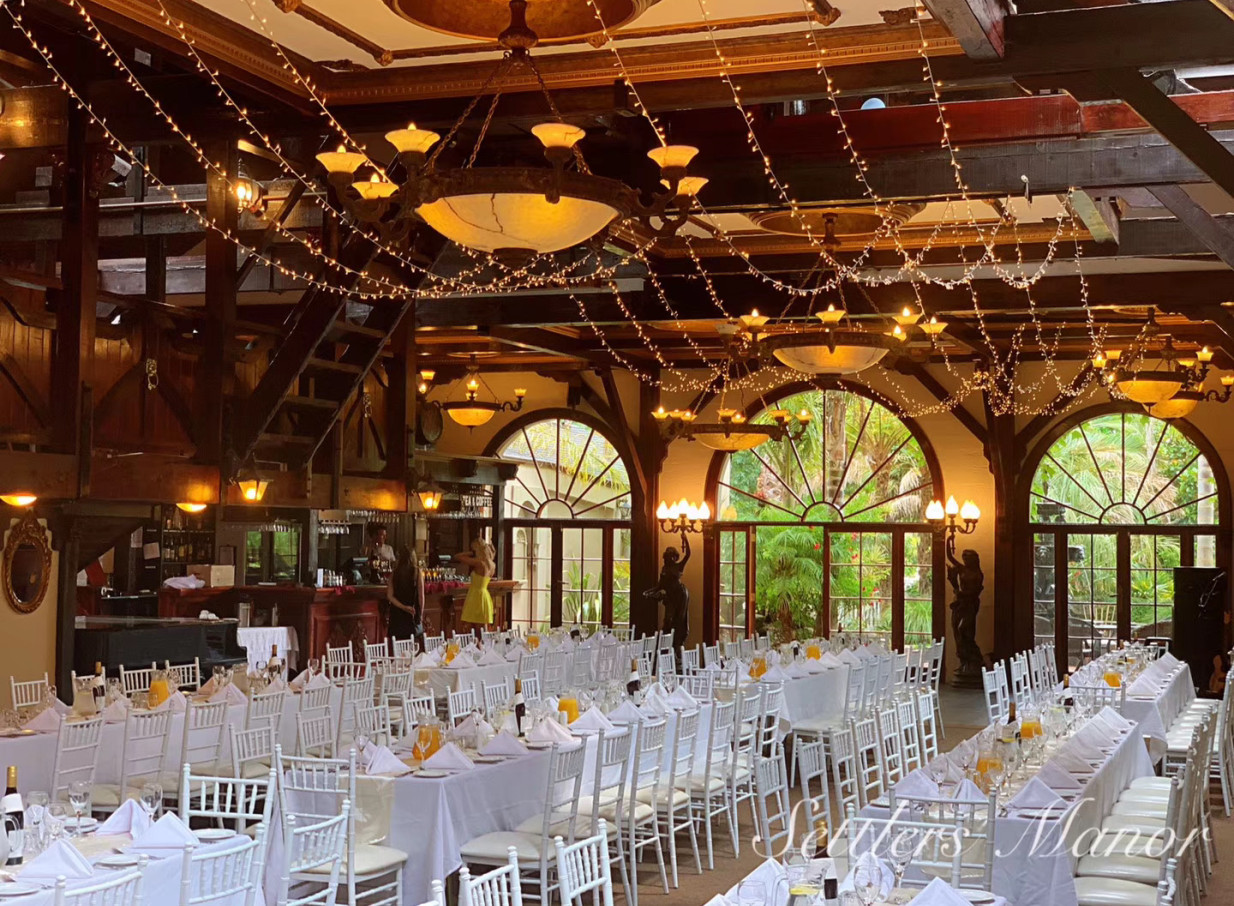 The vintage-style banquet hall at Settlers Country Manor, Auckland, set for an event, features rustic timber beams, arched windows, and fairy lights.