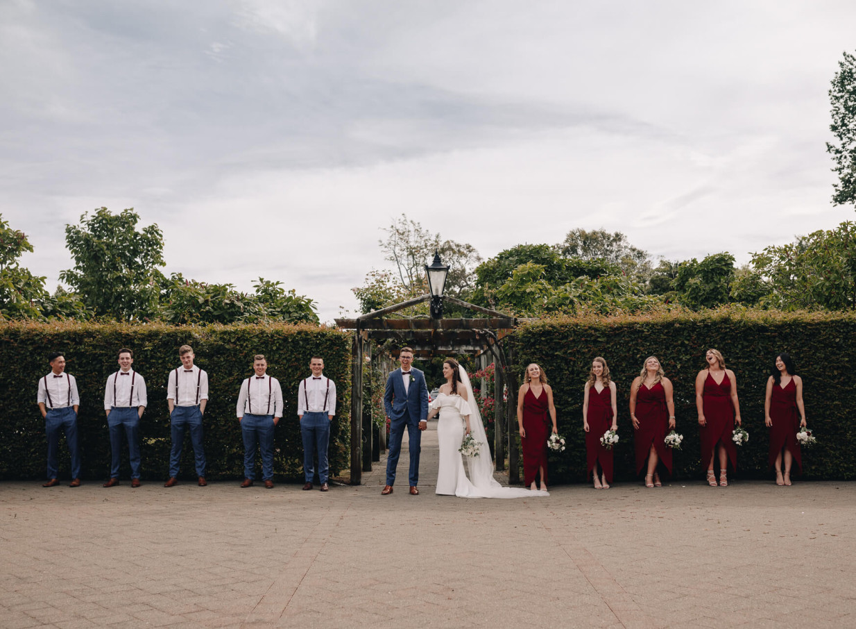 A wedding party smiles near an arbor in the elegant gardens of the Caccia Birch House, a heritage homestead in Manawatū-Whanganui.