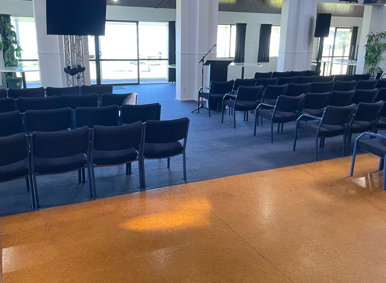 A spacious, light-filled meeting room at the relaxed coastal Tauranga Fish and Dive Club, set for a presentation with rows of chairs and a podium.