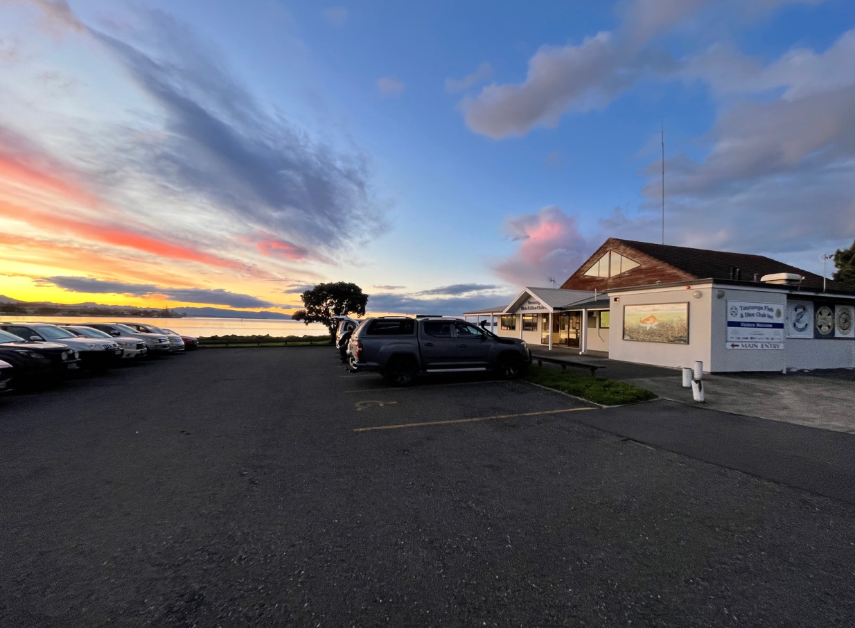 The relaxed coastal exterior of Tauranga Fish and Dive Club with a parking lot and stunning harbour sunset view.