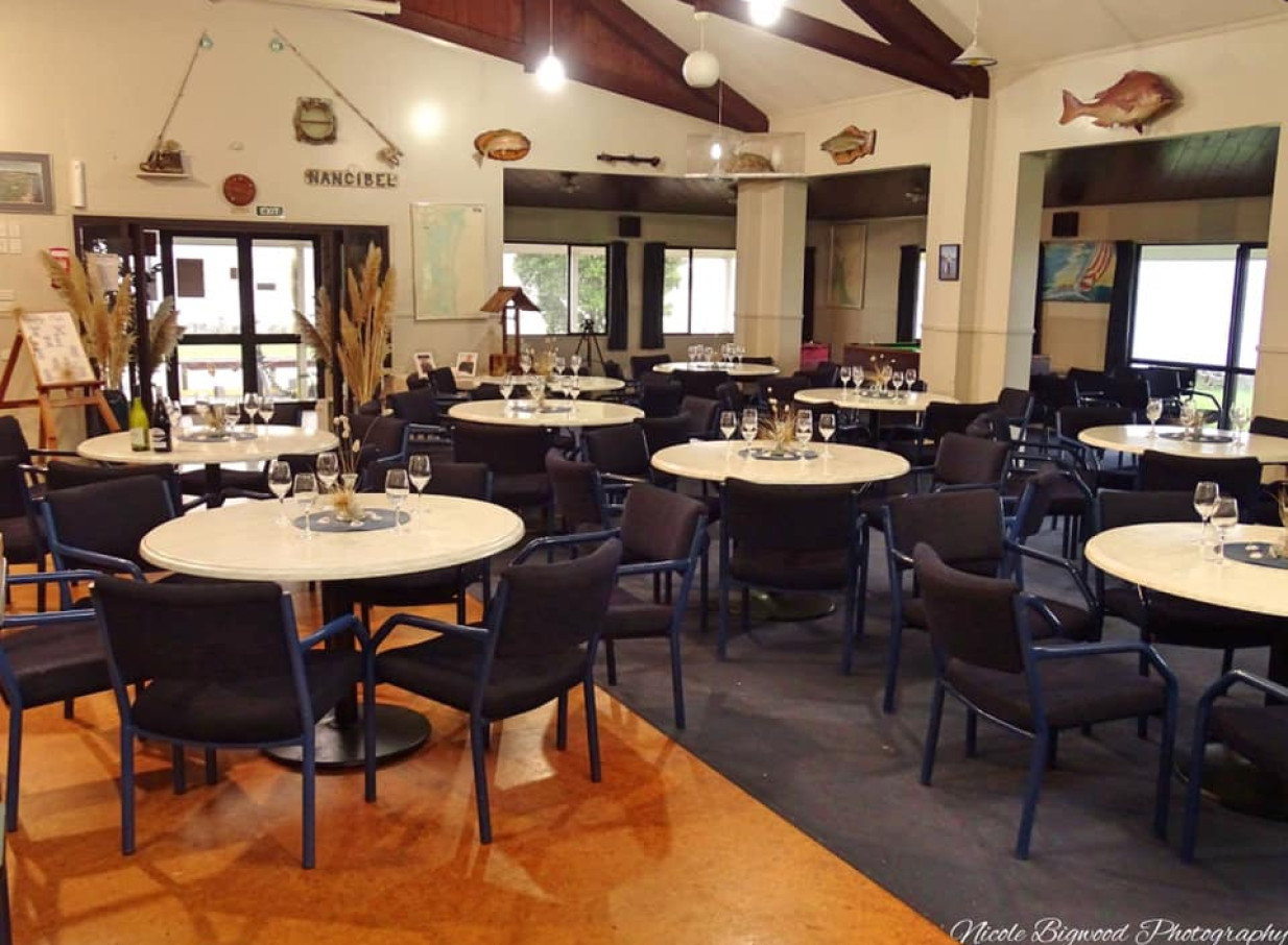 An indoor dining and event space at the Tauranga Fish and Dive Club, featuring a casual seaside style with round tables, wooden beams, and fishing decor.