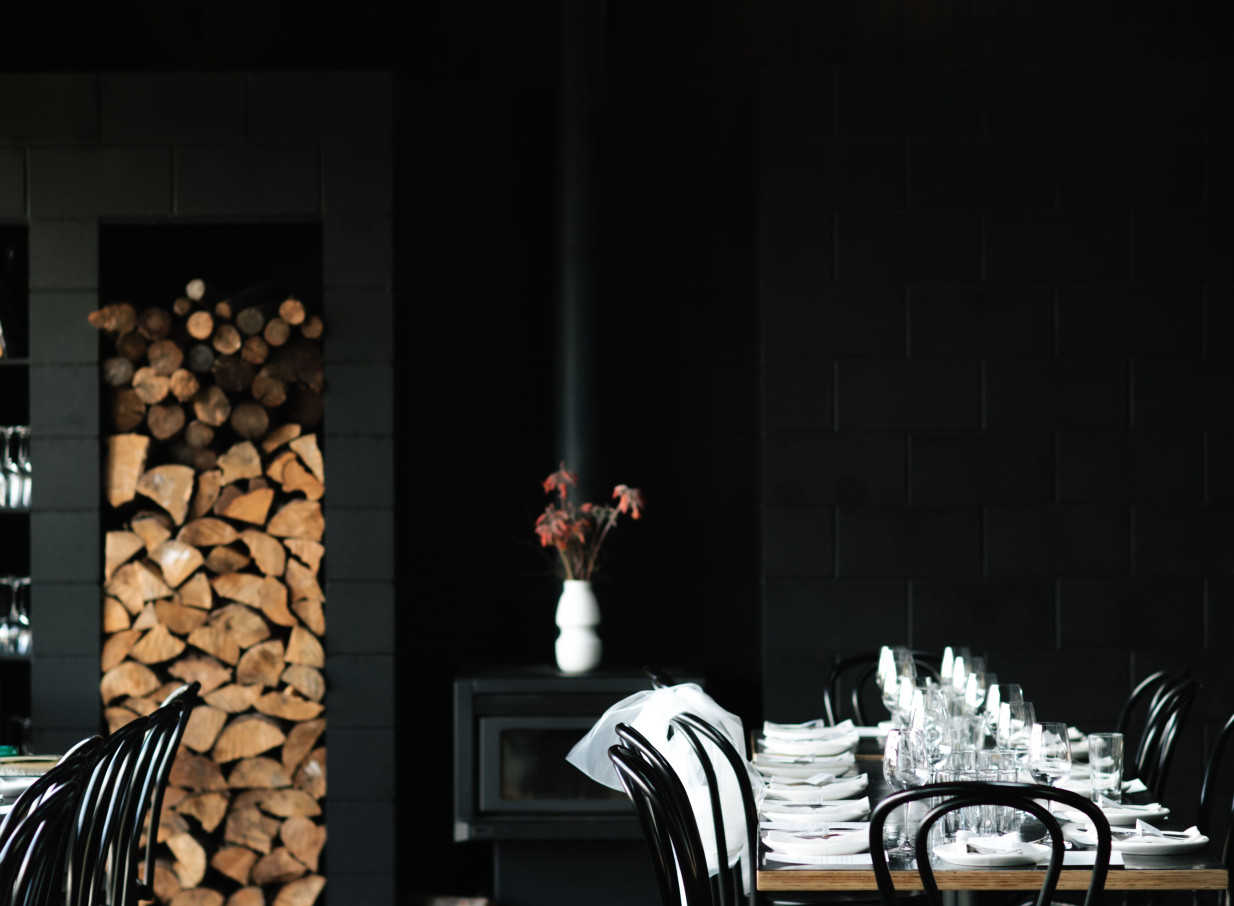 The rustic yet modern dining room at Black Estate Winery in Waipara Valley, near Christchurch, showcases a stacked woodpile and dark decor.
