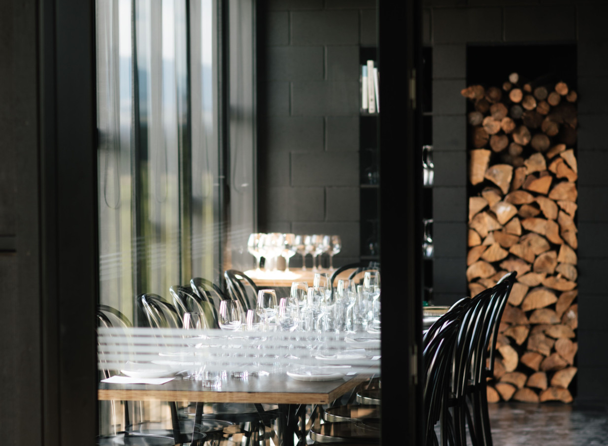 A modern dining room with a set table and a stacked firewood wall at Black Estate Winery, Christchurch, echoing its rustic-chic design.