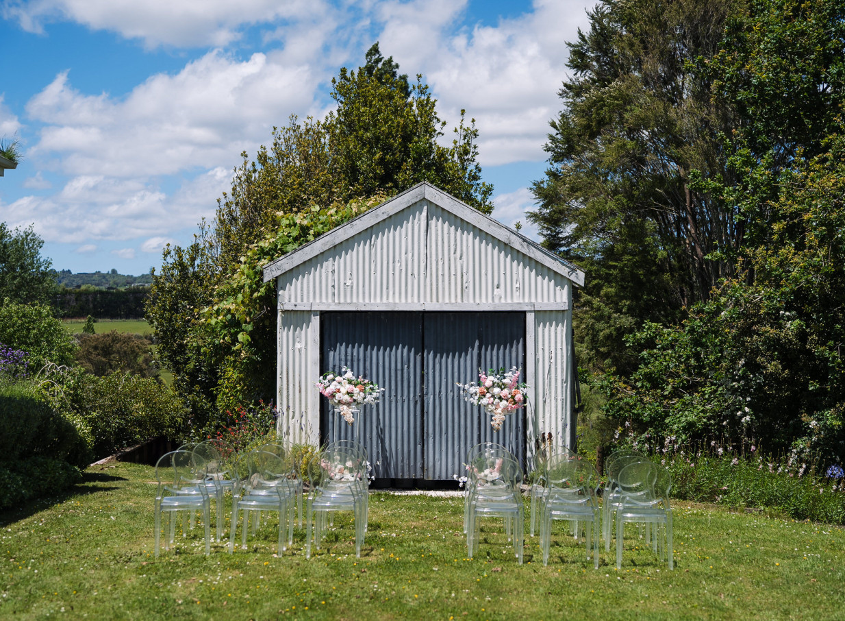 An outdoor ceremony at Auckland's Farm House Cafe, featuring clear chairs and florals in front of a rustic farm building.