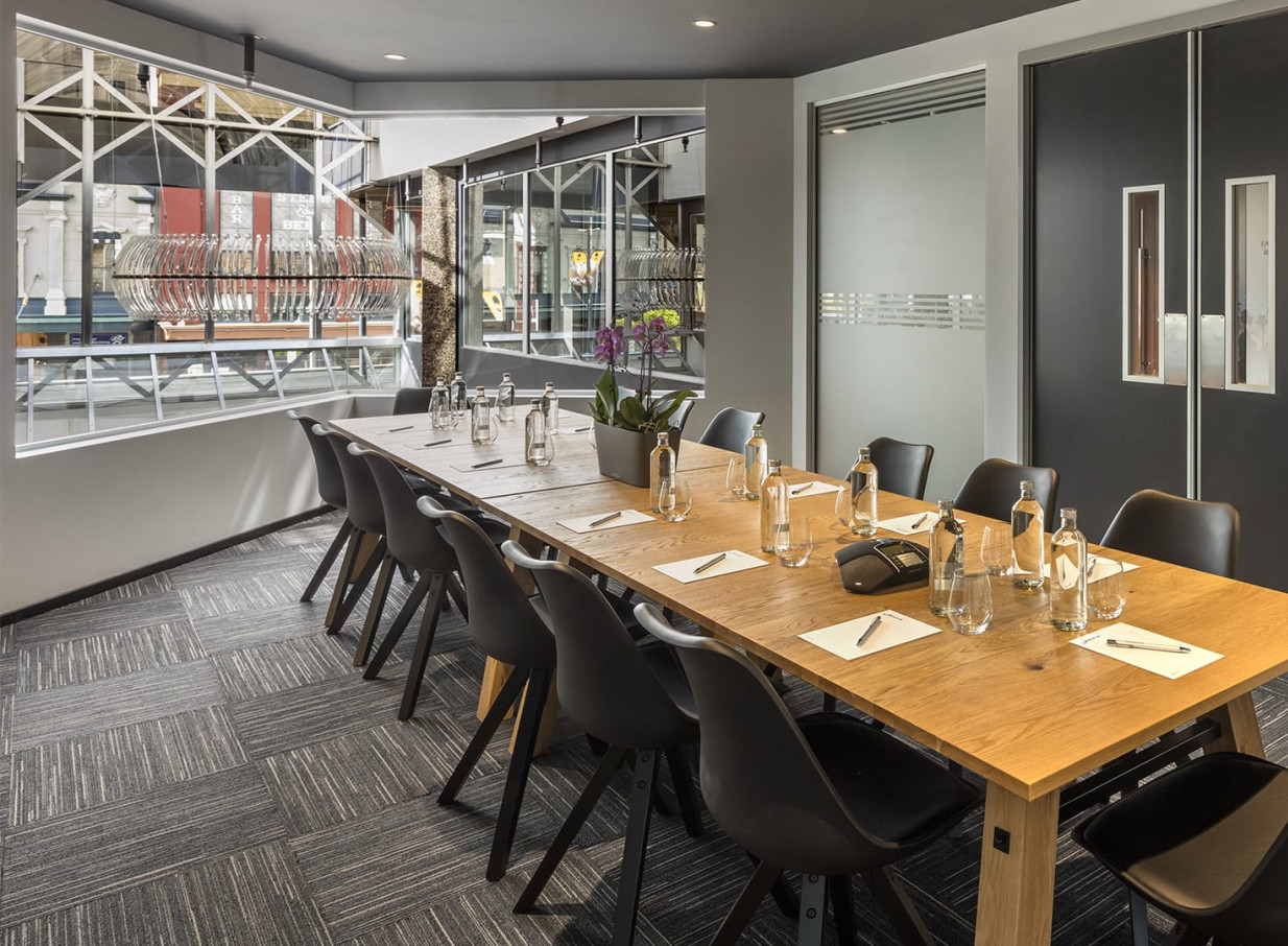 A modern meeting room at the Oaks Wellington Hotel in Wellington, featuring a long wooden table and industrial-chic window details.