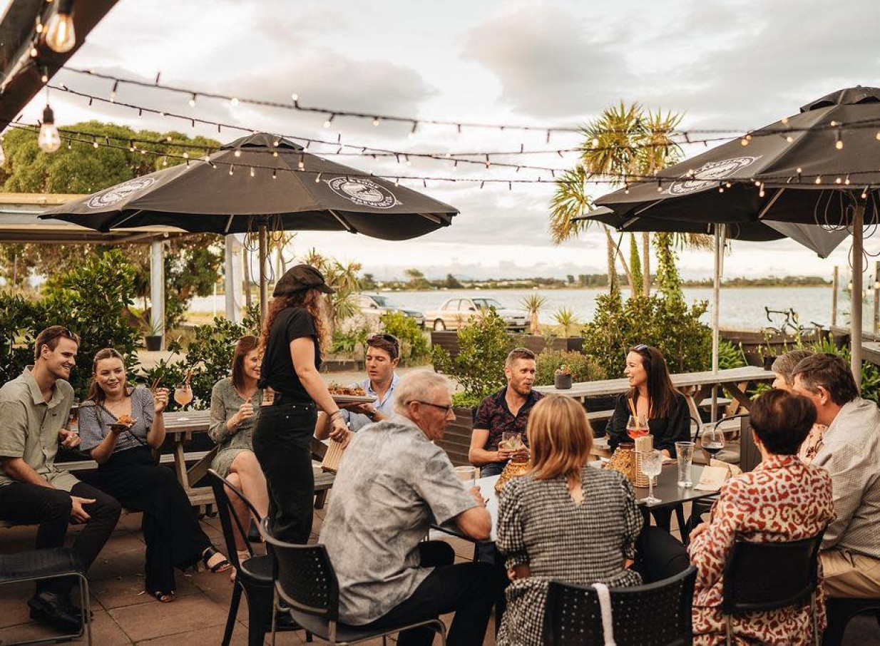 Guests enjoy drinks and shared plates at Evil Genius, a rustic outdoor dining space in Christchurch with estuary views.