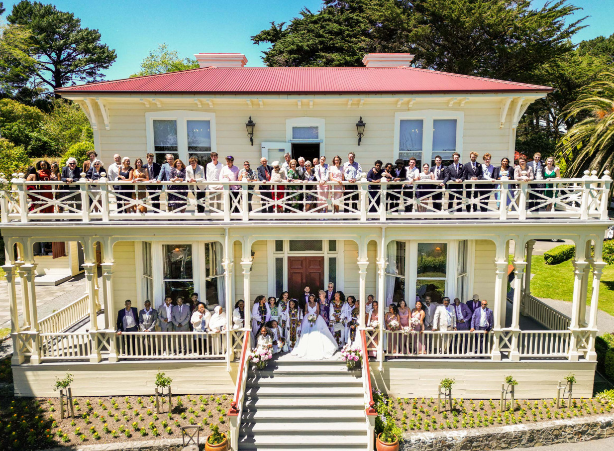 A large wedding party gathers on the wooden verandahs and steps of the historic Victorian Gear Homestead in Porirua, Wellington.