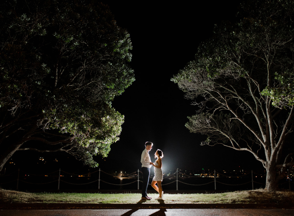 A romantic night shot of a couple under illuminated trees with city lights over the water at Antlia, a heritage waterfront venue in Auckland.