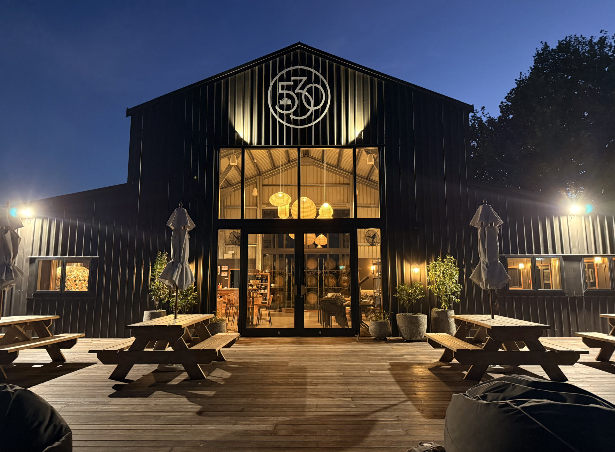 Shed530 Estate, a modern barn-style winery in Hawke's Bay, showing its illuminated interior and outdoor deck at dusk.