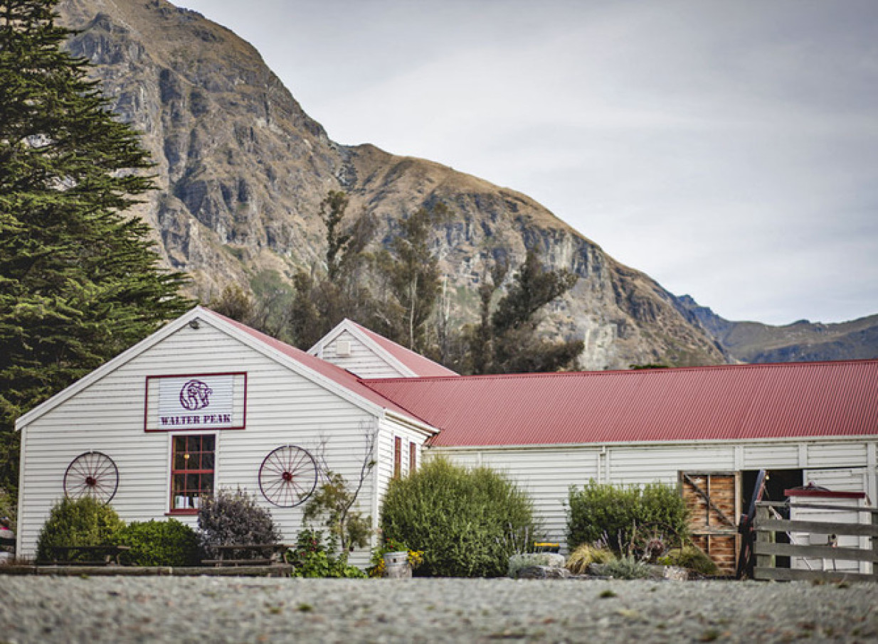 A rustic white farm building with a red roof at Walter Peak, Queenstown, nestled against towering mountains.