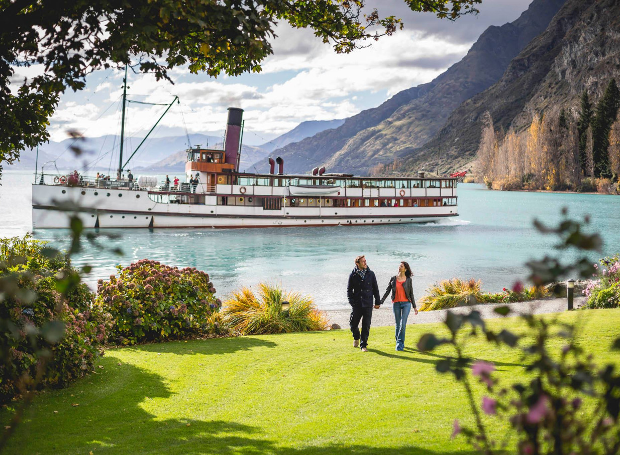 A couple strolls on the manicured lawn of Walter Peak High Country Farm, with the historic TSS Earnslaw vintage steamship cruising on Lake Wakatipu, Queenstown.