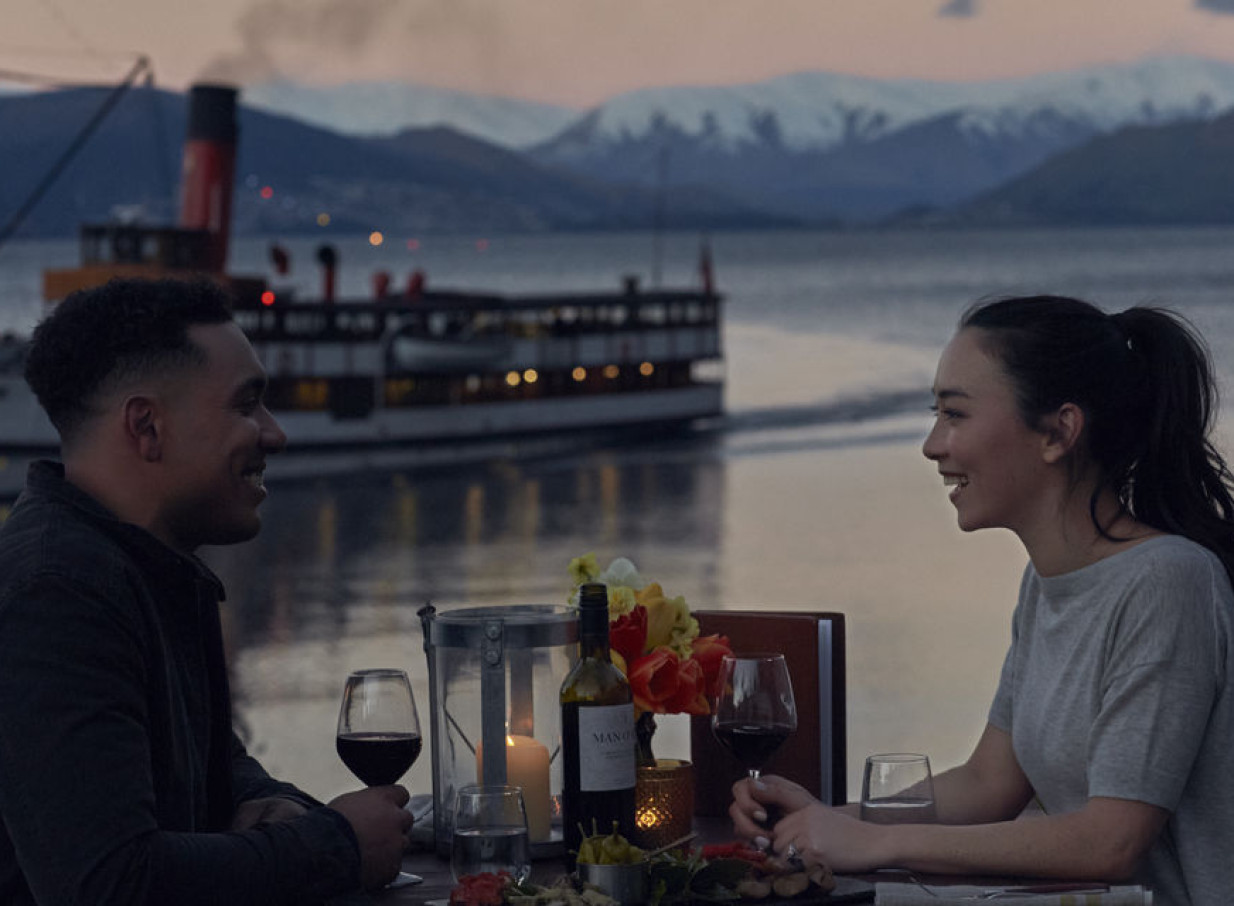 A couple dines al fresco by Lake Wakatipu, with Queenstown's iconic vintage TSS Earnslaw steamship passing by against alpine mountains.