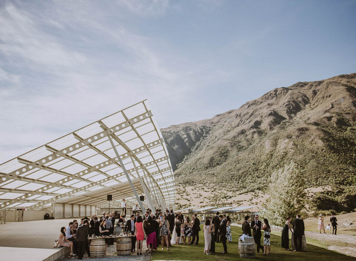 An outdoor reception with guests under Peregrine Winery's falcon-wing inspired 'Under-the-Wing' structure in Queenstown, amidst mountains.