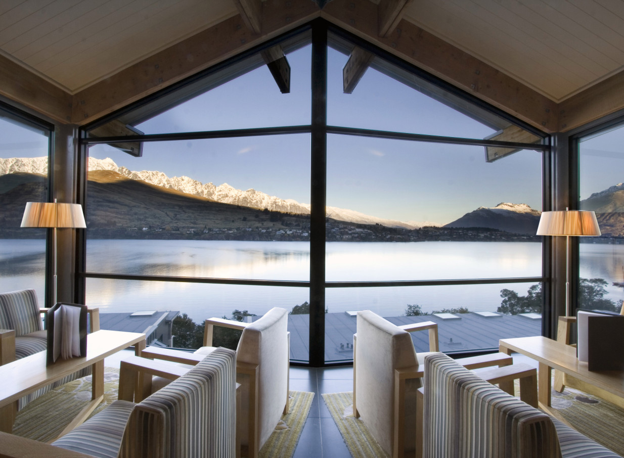 The Rees Hotel, Queenstown, offers a modern lounge with floor-to-ceiling windows framing stunning Lake Wakatipu and mountain views.