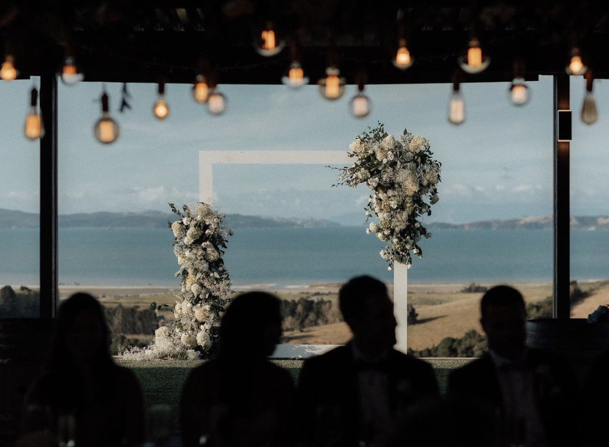 A modern-rustic wedding ceremony at Kauri Bay, Auckland, featuring a floral arch and panoramic coastal views.