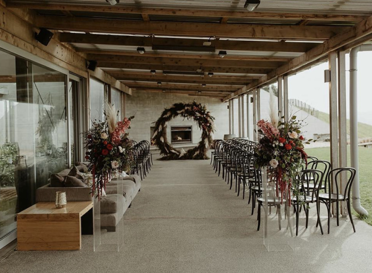 A modern country rustic ceremony space with a wood framework ceiling and floral arch at Kauri Bay in Auckland.
