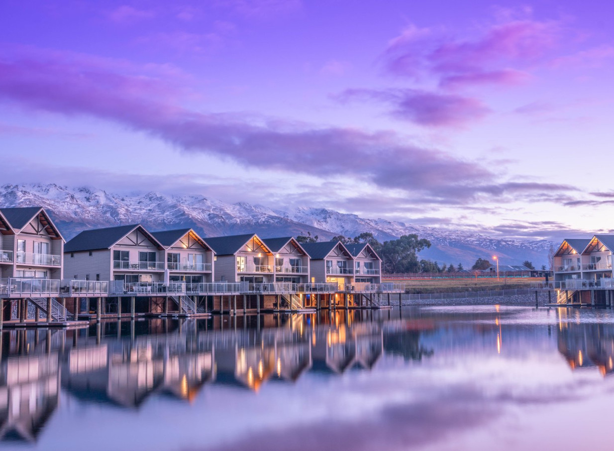 Boatshed-themed villas at Marsden Lake Resort, Central Otago, reflect on Lake Dunstan with snow-capped mountains under a colorful sky.
