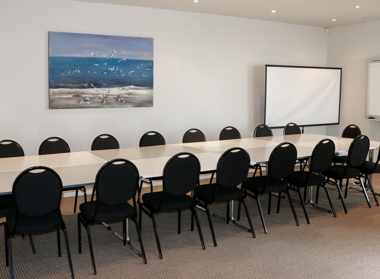 A contemporary meeting room at Butterfly Creek, Auckland, featuring tables, chairs, a projector screen, and seascape art.