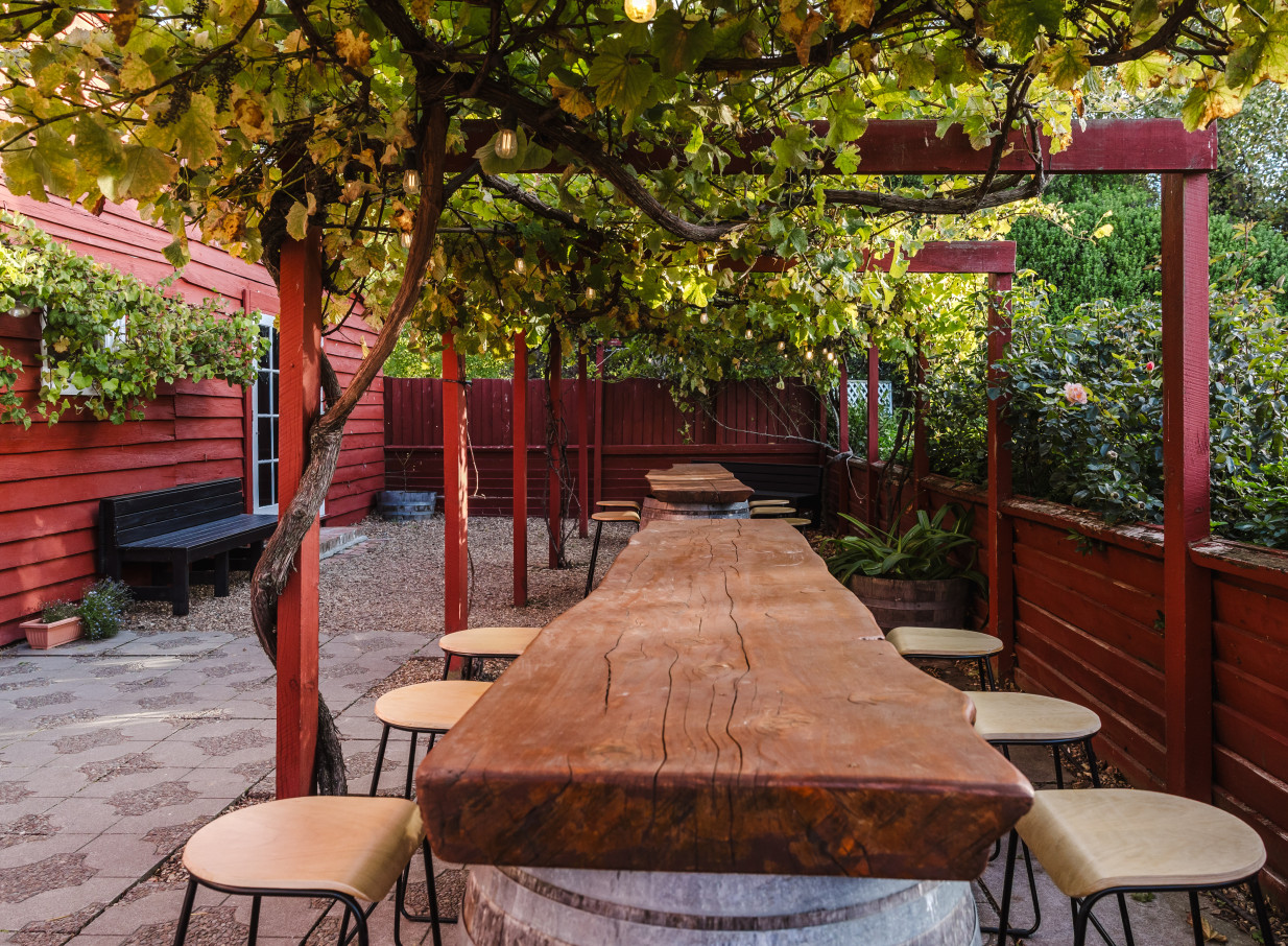 A rustic outdoor dining area with long wooden tables and stools under a vine-covered pergola at Larcomb Vineyard, Christchurch.