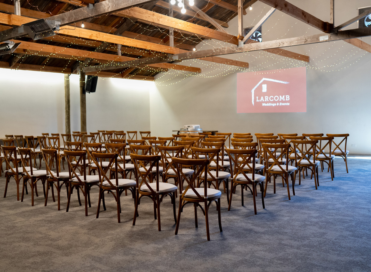 A rustic barn event setup at Larcomb Vineyard, Christchurch, featuring wooden cross-back chairs and exposed beams.