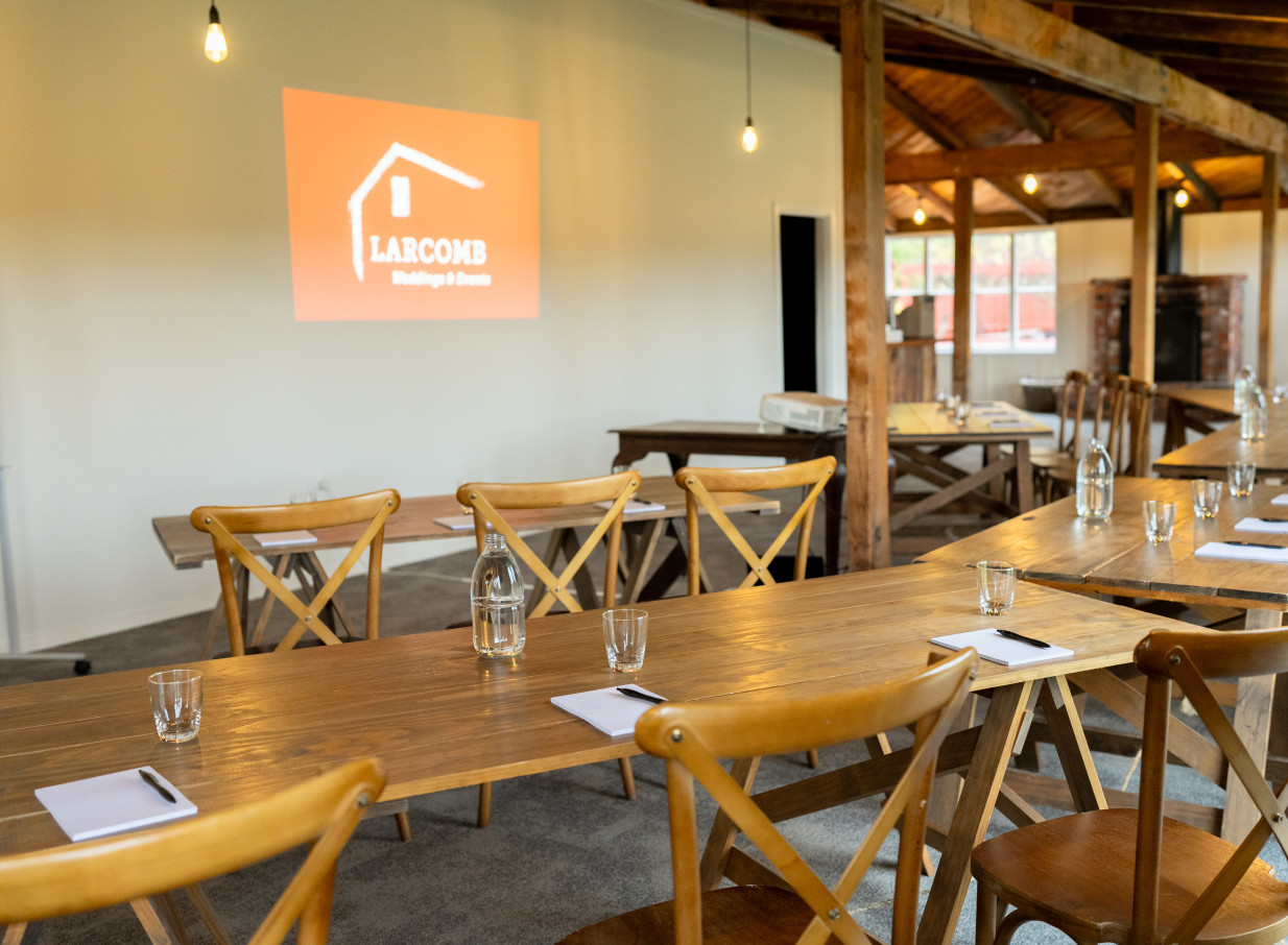 A rustic event room featuring exposed wooden beams and a modern conference setup is visible at Larcomb Vineyard in Christchurch.